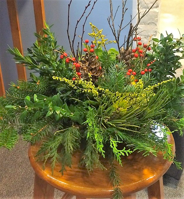 A winter greens centerpiece placed on a wooden stool. The arrangement is made from evergreen branches, holly branches and berries, pine cones, and bare branches.