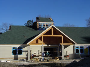 A group of construction workers stands in front of a partially built house with green roofing, blue-trimmed windows, and exposed wooden framing under a clear blue sky.