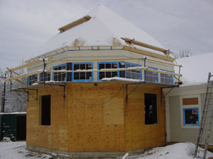 A partially constructed house with a hexagonal shape, wooden exterior, scaffolding, snow on the roof, and snow on the ground. Some windows are installed, and construction materials and a ladder are visible nearby.