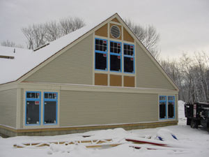 A beige house with blue-trimmed windows and a snow-covered roof stands in a snowy landscape, with bare trees visible in the background. Construction materials are scattered on the snow in the foreground.