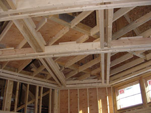 Wooden framework of a house under construction, showing exposed roof trusses, ceiling beams, and wall studs with sunlight coming in through partially installed windows.