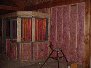 Partially finished room with exposed wooden framing and pink fiberglass insulation installed on the walls. A small unfinished structure with windows is visible, and a metal work stand is in the foreground.