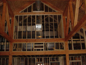 The image shows the interior of a building under construction, featuring exposed wooden beams and metal wall framing with an open loft area and high, sloped ceiling. No drywall or finishing materials are present.