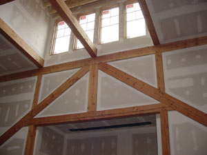 Interior view of a room under construction, showing exposed wooden beams and unfinished drywall with visible seams and patches. Sunlight streams in through four rectangular windows near the ceiling.