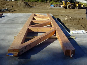 Large wooden roof truss with triangular supports resting on a concrete slab at a construction site, with dirt, machinery, and building materials in the background.