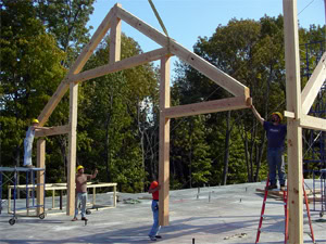 Four construction workers assemble a wooden frame structure on a concrete foundation outdoors, surrounded by trees. Two workers stand on ladders, while two others work on the ground, all wearing safety gear.