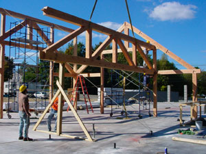 Construction workers guide a large wooden roof truss being lifted into place by a crane at a building site. Scaffolding, ladders, and tools are visible, with a clear blue sky in the background.