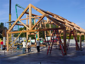 Workers wearing hard hats stand near the wooden frame of a building under construction, with ladders and construction equipment visible on a sunny day.
