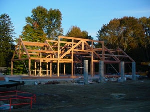 Wooden framing of a building under construction, surrounded by trees and construction materials on the ground, in an outdoor setting at sunset.
