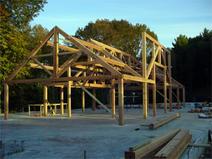 A wooden frame structure of a building under construction stands on a concrete foundation, surrounded by trees and scattered building materials under a clear sky.