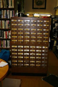 A wooden library card catalog with multiple small drawers and brass label holders stands in a room lined with bookshelves.