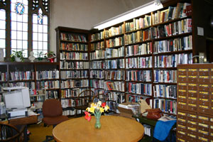 A cozy library corner with tall shelves full of books, a wooden table with a vase of flowers, a few chairs, a brown card catalog, and a computer by the window with stained glass.