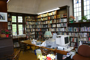 A cozy library room with tall bookshelves filled with books, wooden chairs around a large table, and a desk with an old computer, printer, and a lamp. Sunlight shines through large windows.