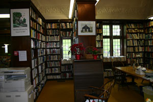 A cozy library room with tall bookshelves filled with books, a wooden table with chairs, potted plants on a shelf, large windows letting in natural light, and a printer in the corner.