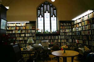 A cozy library room with tall bookshelves filled with books, a large arched window with panes letting in light, and a round table with flowers in a vase in the center.