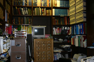 A cluttered office with shelves full of books, binders, and files; a desk with a vintage computer monitor, filing cabinets, and stacks of papers and folders scattered around.