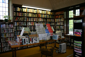 A cozy library room with tall bookshelves filled with books, wooden tables and chairs, display racks with pamphlets, and computers by the windows letting in natural light.
