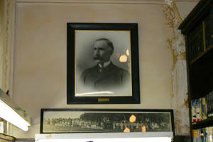 Framed black-and-white portrait of a man with a mustache hangs on a wall above a wide, panoramic photo; parts of the wall are stained or peeling, and bookshelves are visible on the right.