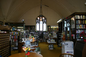 A cozy library interior with arched ceilings, shelves filled with books, a central chandelier, computers, printers, and a table with a vase of flowers in the foreground.