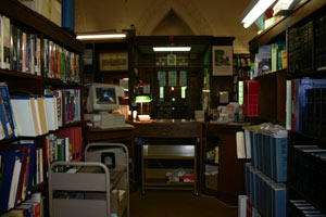 A dimly lit library workspace with bookshelves on both sides, a desk with a computer and lamp in the center, and a book cart in the aisle. A wooden door with glass panes is visible at the back.