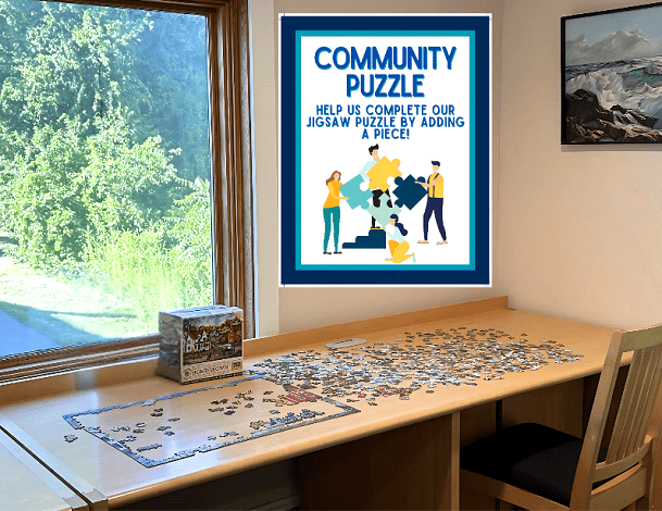 A jigsaw puzzle in progress sits on a wooden table by a window with a scenic view. A sign on the wall reads, “Community Puzzle. Help us complete our jigsaw puzzle by adding a piece!”.