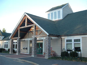 A one-story public library with a green roof, stone pillars, and large windows at the entrance. Bike racks and a bench are outside, and sunlight shines on the building.