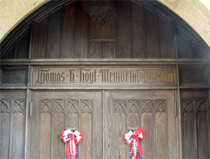 Wooden double doors with arched panels and decorative carvings. Above the doors, a sign reads Thomas T. Hoyt Memorial Library. Two red, white, and blue ribbon bows are attached to the doors.