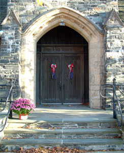 Stone church entrance with arched doorway, double wooden doors decorated with red and white bows. A potted plant with pink flowers sits on the steps leading up to the entrance.
