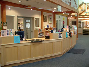 A spacious library front desk with light wood counters, computers, books, and brochures. Shelves and office spaces are visible behind the desk, with bright lighting and large windows in the background.