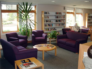 A cozy library reading area with purple armchairs and a sofa around a round wooden table. Bookshelves line the walls, and a person sits reading at a table near large windows letting in natural light.