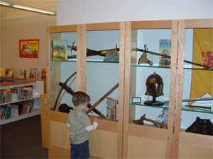 A young boy stands in front of a museum display case containing historical weapons, a helmet, and various artifacts. Bookshelves and colorful posters are visible in the background.