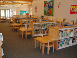 A childrens library section with low bookshelves filled with books, small wooden tables and chairs, and colorful artwork on the walls. The space is well-lit and inviting for young readers.