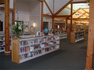 A spacious library interior with wooden beams, bookshelves filled with books, a globe on one shelf, a vase of flowers on the counter, and natural light coming through large windows.