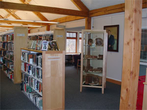 Bookshelves filled with books line a library interior with wooden beams. A display case containing artifacts stands in the center, and a portrait hangs on the wall in the background near a window.
