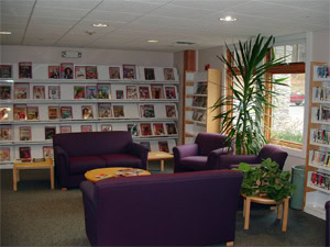 A cozy library reading area with purple sofas and armchairs, small tables with plants, and bookshelves lining the walls filled with magazines and books. A large window lets in natural light.