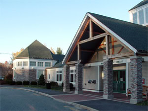 A modern library building with stone columns and large windows, featuring a peaked roof and a covered entrance, sits next to a similarly styled round building. Trees and clear sky are visible in the background.