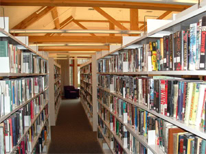 Bookshelves filled with books line both sides of a library aisle, with a wooden beam ceiling above and a window at the end letting in natural light.