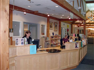 A library front desk with two people working at computers; bookshelves, office windows, and informational flyers are visible in the background. The area appears well-lit and organized.