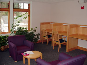 A cozy study room with two wooden study carrels and chairs, two purple armchairs, a small round table with a notebook and pen, large windows, and green potted plants.