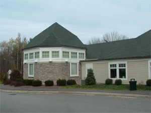 A beige building with a green, pyramid-shaped roof and many windows, surrounded by bushes and trees, sits under a cloudy sky next to a paved road.