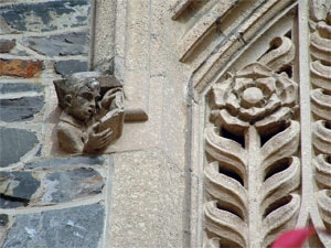 Stone carving of a small figure reading a book on the wall of a Gothic-style building, next to an ornate floral stone relief.