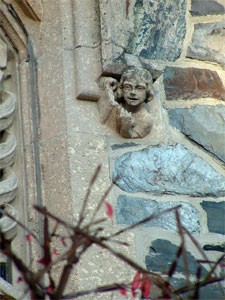 A small stone cherub sculpture is embedded in a building’s corner, with its head and arm visible. The wall is made of rough stone and mortar, and branches with red buds are in the foreground.
