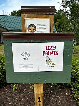 A green outdoor story display features the cover of the children’s book Izzy Paints, showing a cartoon animal painting. Another sign with text and a logo is posted above it. Trees and bushes are in the background.