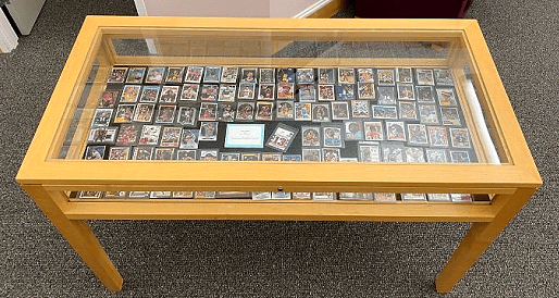 A glass display case with wooden legs showcases a large collection of trading cards arranged in rows on a dark surface, set on a carpeted floor.