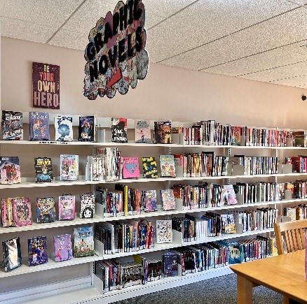 A library wall displays graphic novels and comics on white shelves, with various colorful book covers visible. A “GRAPHIC NOVELS” sign hangs above and a poster reads “BE YOUR OWN HERO.” Part of a wooden chair and table are in view.