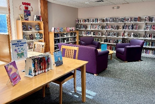 A cozy library area with purple armchairs, bookshelves filled with books, and a wooden table displaying middle grade fiction novels near a sunlit window.