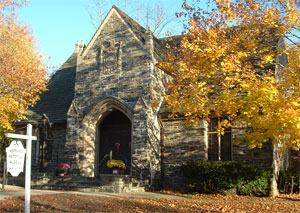 A small stone church with a pointed arch entrance, surrounded by trees with golden autumn leaves. A white sign is visible in the foreground near the path to the entrance.