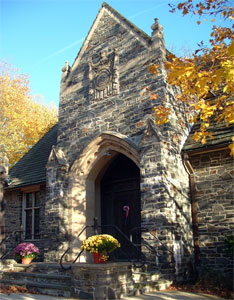 Stone church entrance with an arched doorway, ornate stonework, and a crest above the door. Autumn trees with yellow leaves surround the building, and potted flowers sit by the entrance steps.
